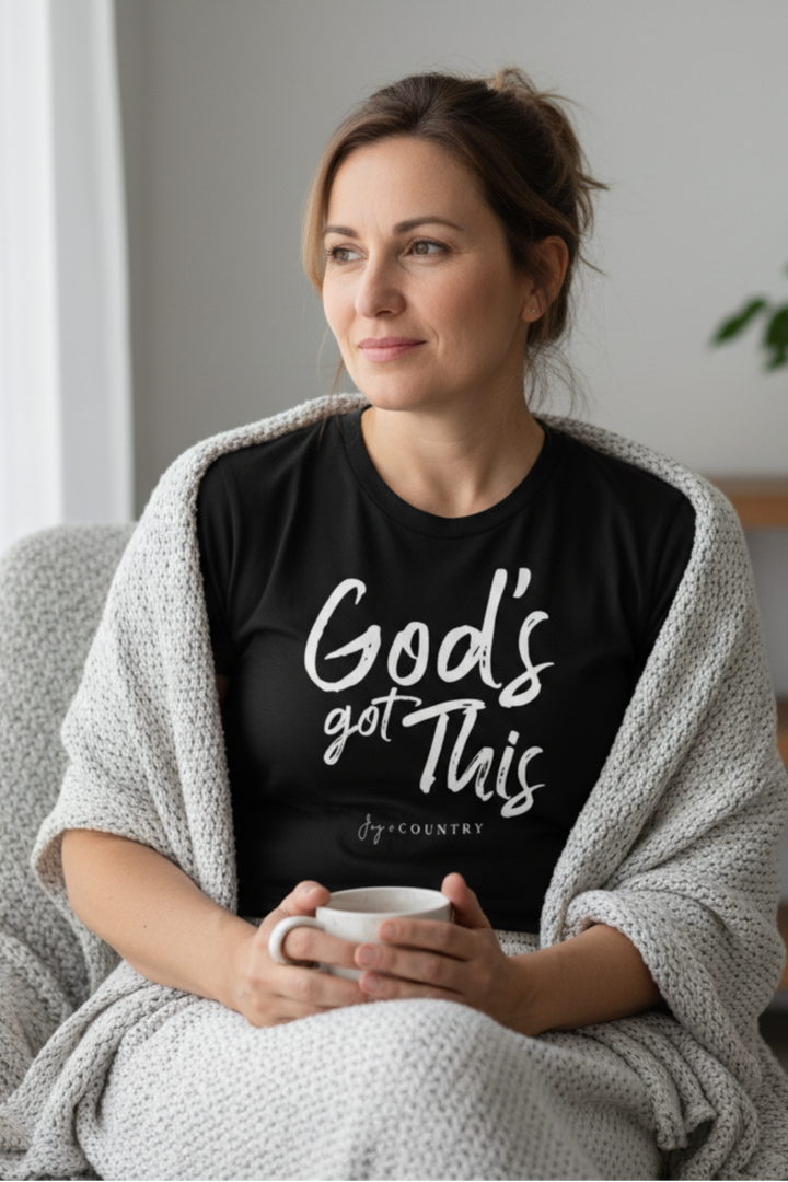 Woman wearing a black t-shirt with 'God's got this' text, holding a mug in a cozy indoor setting.