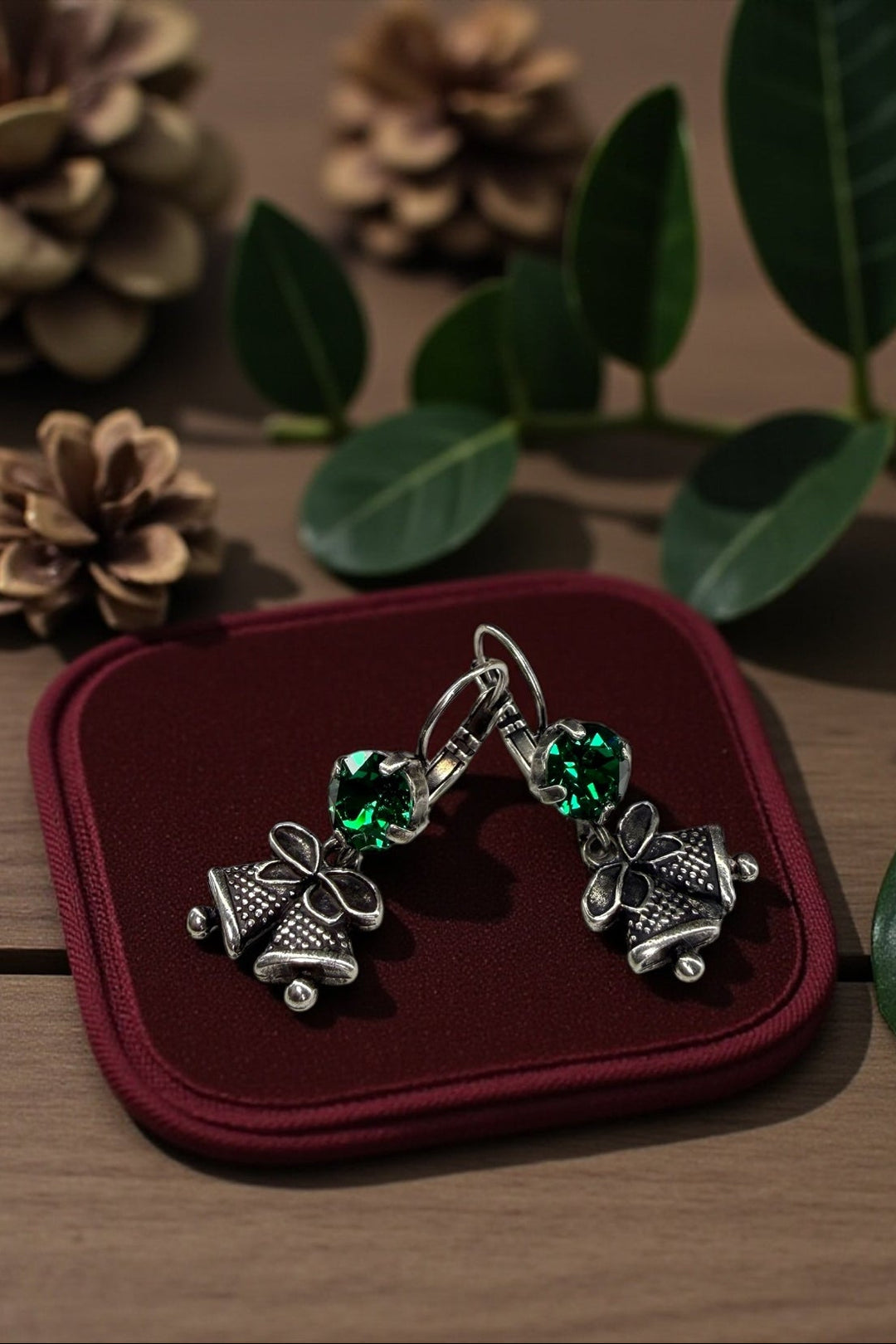 Silver Christmas bell earrings with green crystals on a red cushion, surrounded by decorative elements.