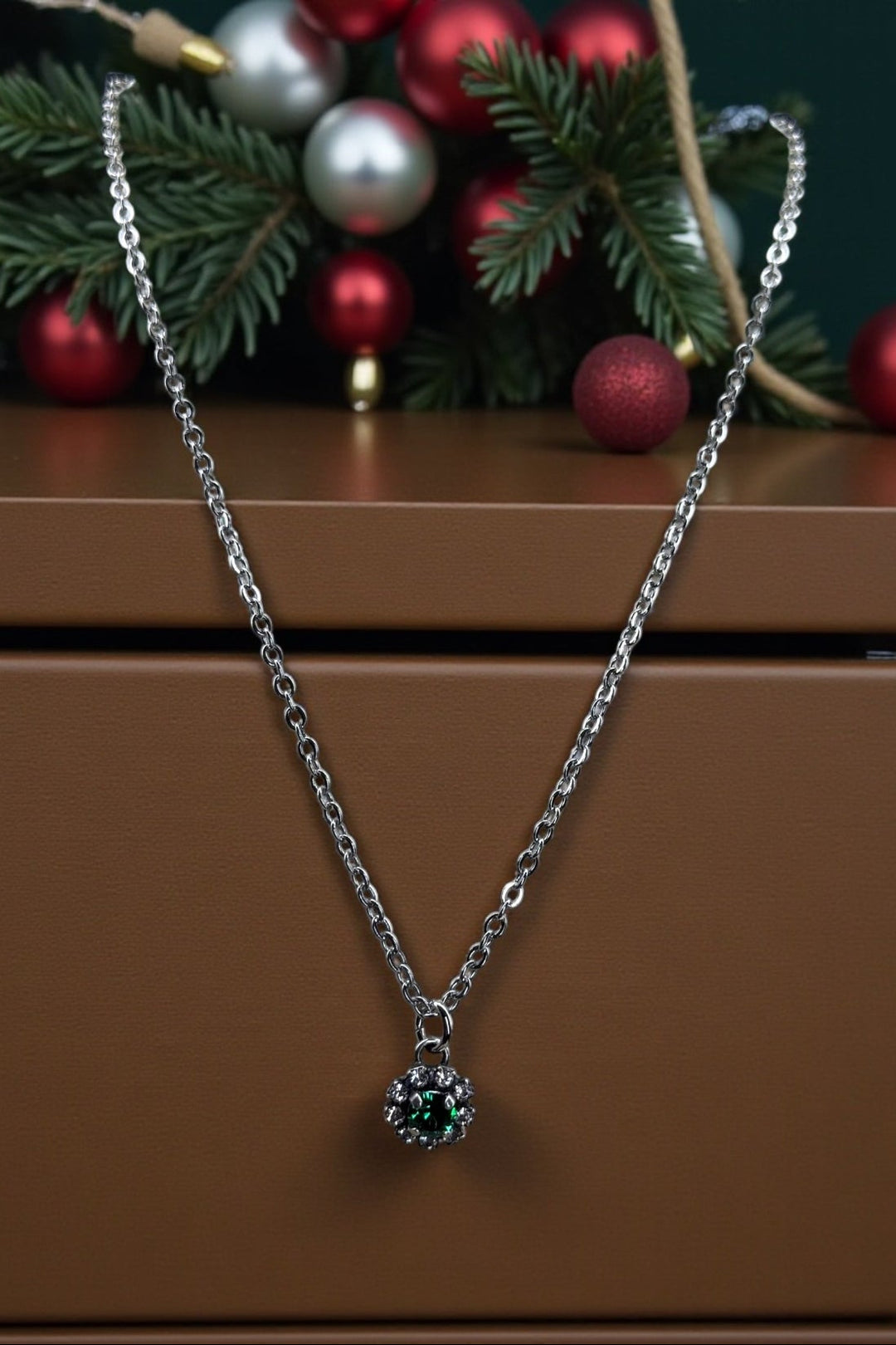 Silver necklace with a green crystal pendant on a brown surface with Christmas decorations in the background.
