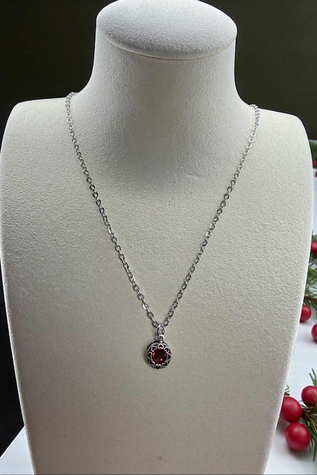 Silver necklace with a red crystal pendant with a silver heart surround, on a mannequin bust, with berries and greenery in the background.