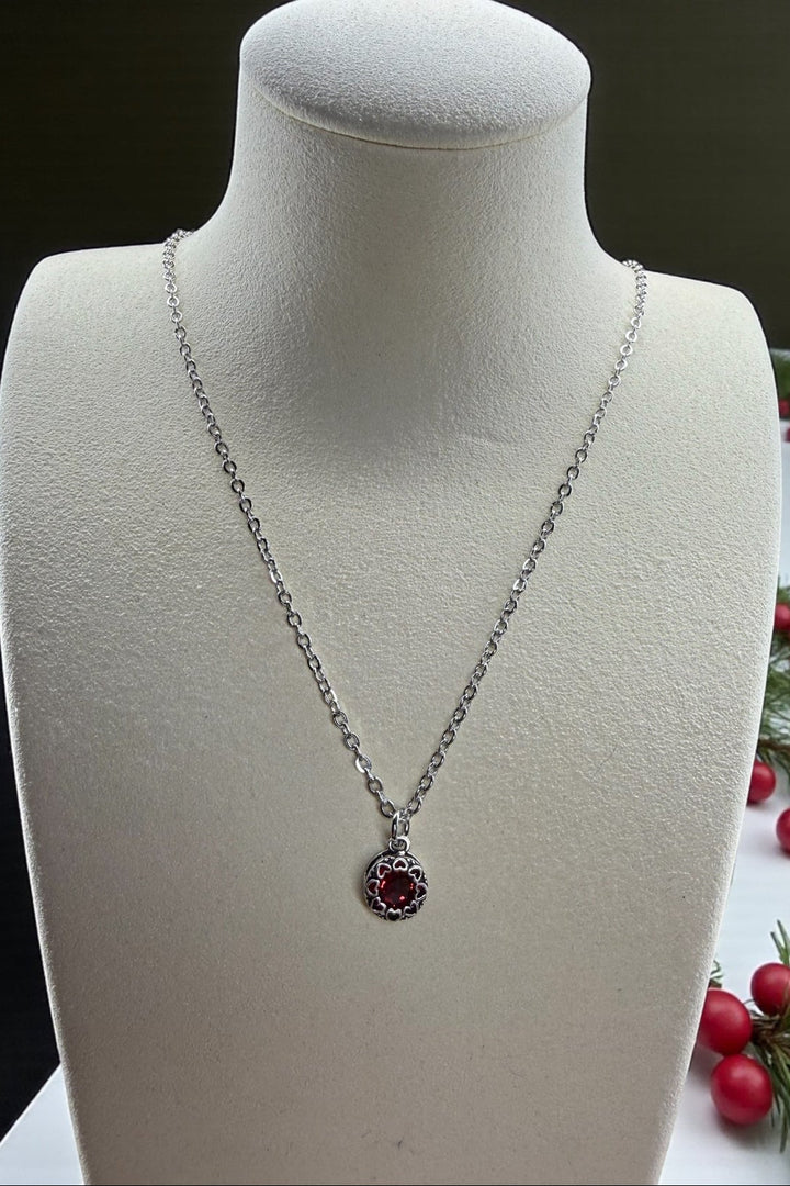 Silver necklace with a red crystal pendant with a silver heart surround, on a mannequin bust, with berries and greenery in the background.